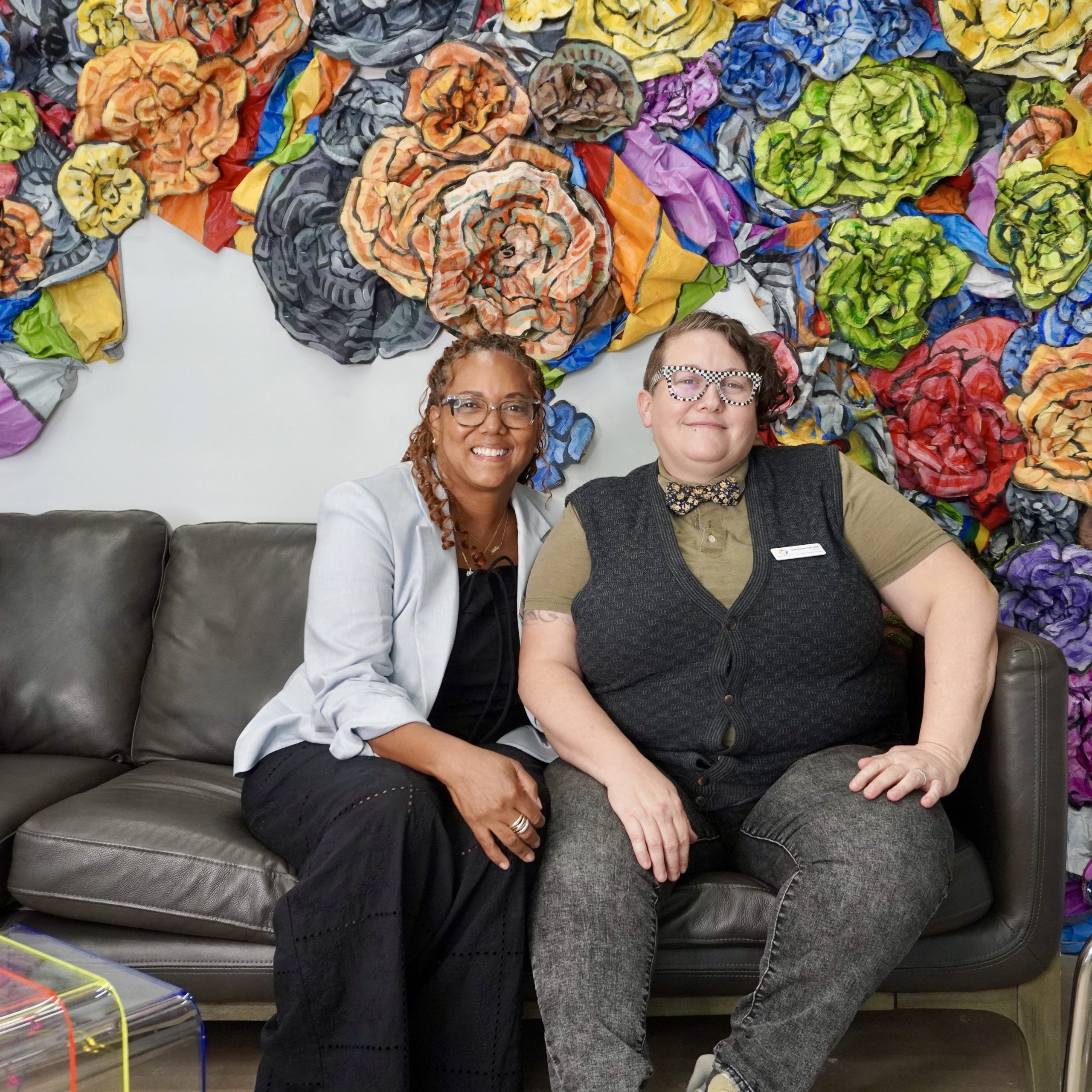 Two people seated in a community arts space with colorful floral artwork behind them.